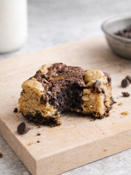 Brookie maison partiellement croqué sur planche en bois, mélange de brownie fondant et cookie doré, avec éclats de chocolat et verre de lait en arrière‑plan.