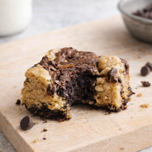 Brookie maison partiellement croqué sur planche en bois, mélange de brownie fondant et cookie doré, avec éclats de chocolat et verre de lait en arrière‑plan.