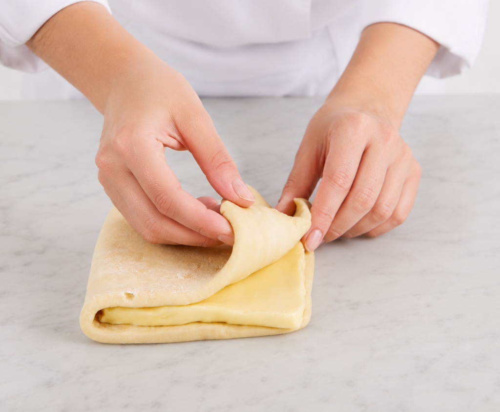 Mains de femme pliant une pâte feuilletée sur une table en marbre gris très clair dans une cuisine lumineuse. Gros plan sur des mains de femme en veste de pâtissière pliant une pâte feuilletée contenant une couche visible de beurre. La scène est photographiée sous un angle rapproché et frontal, avec une lumière douce et naturelle. Le plan de travail en marbre gris très clair et l’arrière-plan épuré mettent en valeur la texture de la pâte et le geste artisanal de préparation pâtissière.