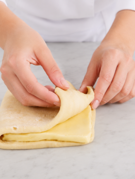 Mains de femme pliant une pâte feuilletée sur une table en marbre gris très clair dans une cuisine lumineuse. Gros plan sur des mains de femme en veste de pâtissière pliant une pâte feuilletée contenant une couche visible de beurre. La scène est photographiée sous un angle rapproché et frontal, avec une lumière douce et naturelle. Le plan de travail en marbre gris très clair et l’arrière-plan épuré mettent en valeur la texture de la pâte et le geste artisanal de préparation pâtissière.
