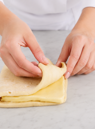 Mains de femme pliant une pĂąte feuilletĂ©e sur une table en marbre gris trĂšs clair dans une cuisine lumineuse. Gros plan sur des mains de femme en veste de pĂątissiĂšre pliant une pĂąte feuilletĂ©e contenant une couche visible de beurre. La scĂšne est photographiĂ©e sous un angle rapprochĂ© et frontal, avec une lumiĂšre douce et naturelle. Le plan de travail en marbre gris trĂšs clair et lâarriĂšre-plan Ă©purĂ© mettent en valeur la texture de la pĂąte et le geste artisanal de prĂ©paration pĂątissiĂšre.