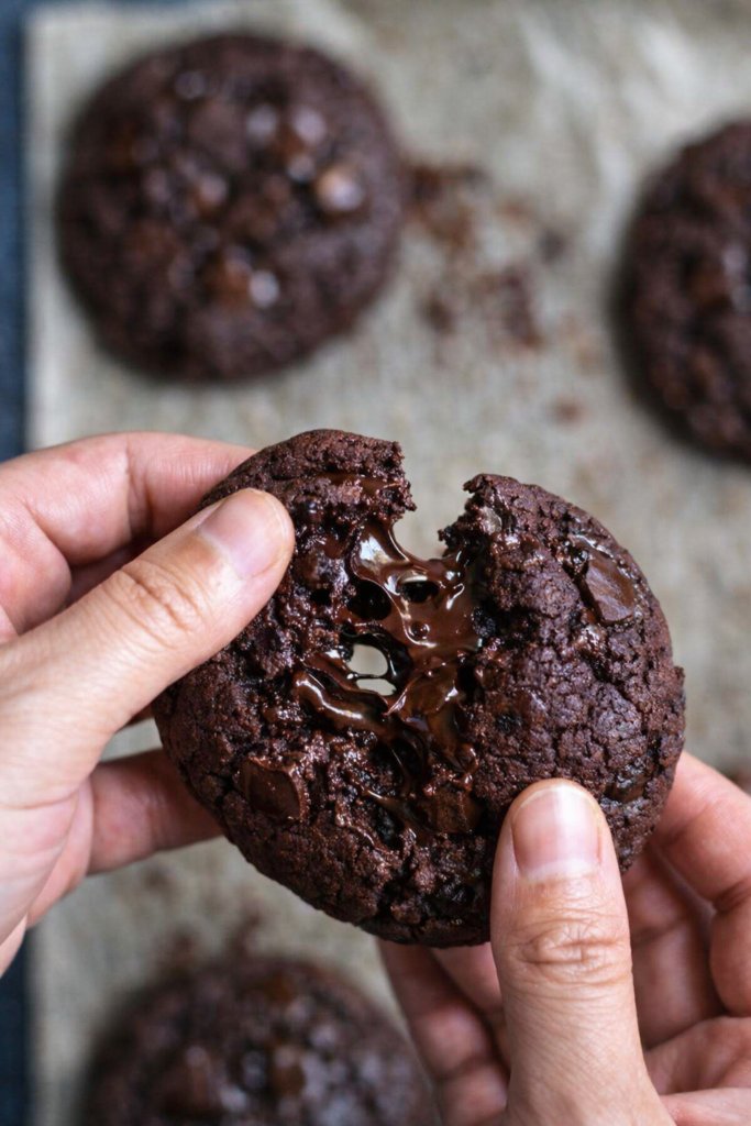 Cookie double chocolat cassé en deux, cœur fondant et morceaux de chocolat noir fondus, photo culinaire rustique sur fond en bois.