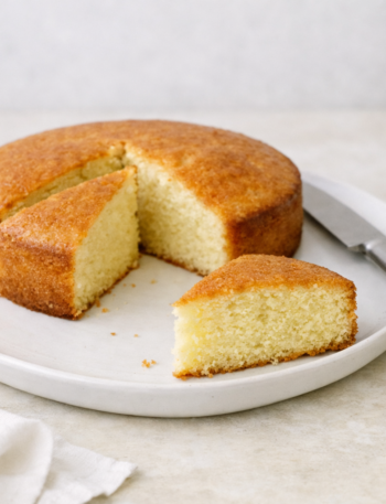 Gâteau au yaourt doré et moelleux sur une assiette blanche, avec une part découpée, éclairage naturel et décor minimaliste, style culinaire épuré et professionnel.