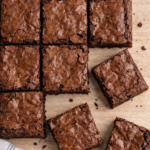 Carrés de brownies fondants au chocolat disposés sur une table en bois clair, avec un torchon blanc à liserets bleus visible en bas de l’image.