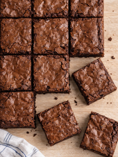 Carrés de brownies fondants au chocolat disposés sur une table en bois clair, avec un torchon blanc à liserets bleus visible en bas de l’image.