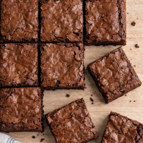 Carrés de brownies fondants au chocolat disposés sur une table en bois clair, avec un torchon blanc à liserets bleus visible en bas de l’image.