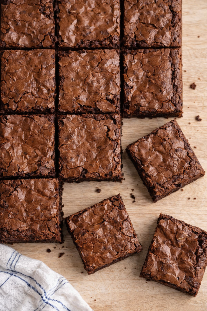 Carrés de brownies fondants au chocolat disposés sur une table en bois clair, avec un torchon blanc à liserets bleus visible en bas de l’image.