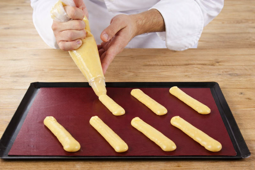 Pâtissier en veste blanche pochant de la pâte à choux sur une plaque noire recouverte d’un tapis de cuisson rouge bordeaux, sur une table en bois clair.
