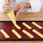Pâtissier en veste blanche pochant de la pâte à choux sur une plaque noire recouverte d’un tapis de cuisson rouge bordeaux, sur une table en bois clair.