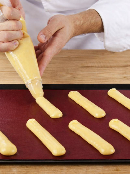 Pâtissier en veste blanche pochant de la pâte à choux sur une plaque noire recouverte d’un tapis de cuisson rouge bordeaux, sur une table en bois clair.