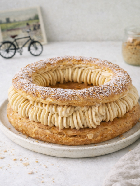 Recette de Paris‑Brest traditionnelle maison sur table blanche, dessert français en pâte à choux dorée garni de crème pralinée, lumière naturelle du jour, décor minimaliste avec mini vélo vintage flou en arrière‑plan.