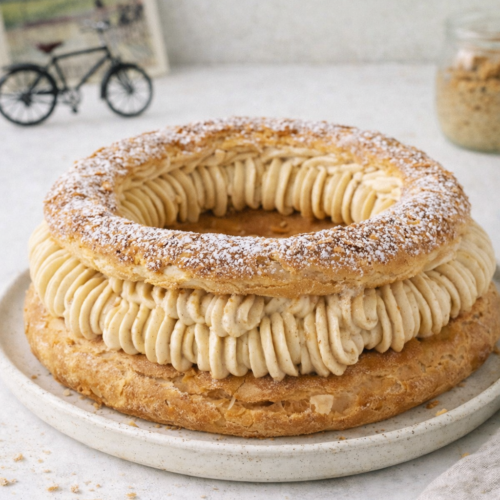 Paris‑Brest traditionnel praliné maison sur table blanche, dessert français en pâte à choux dorée garni de crème pralinée, lumière naturelle du jour, décor minimaliste avec mini vélo vintage flou en arrière‑plan.