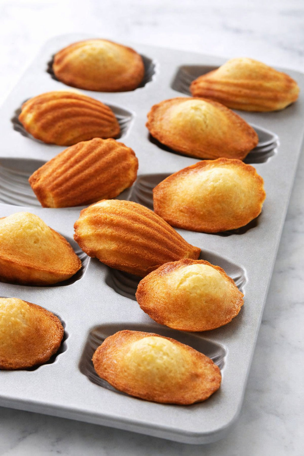 Madeleines maison dorées dans un moule gris clair, éclairées par une lumière naturelle, ambiance gourmande et artisanale.
