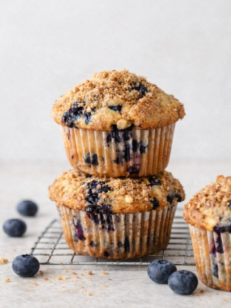Muffins aux myrtilles dorés avec topping croustillant, posés sur fond clair, entourés de quelques myrtilles fraîches, lumière naturelle douce.