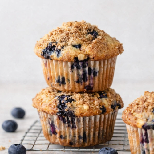 Muffins aux myrtilles dorés avec topping croustillant, posés sur fond clair, entourés de quelques myrtilles fraîches, lumière naturelle douce.