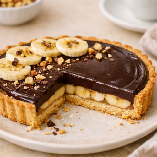 Tarte choco‑banane fine présentée sur une assiette beige, avec une part manquante révélant les couches de pâte sablée, banane et ganache chocolat brillante, dans une lumière naturelle et un décor minimaliste.