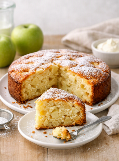Gâteau aux pommes moelleux photographié en lumière naturelle, posé sur une table en bois claire avec une part découpée et saupoudrée de sucre glace.