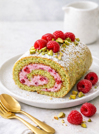 Gâteau roulé à la pistache garni de crème framboise, présenté sur une assiette en grès claire avec couverts dorés, décoré de framboises fraîches et éclats de pistaches.