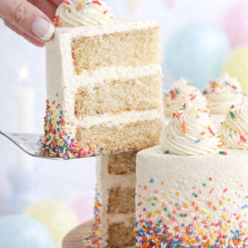 Gâteau vanille à trois étages recouvert d’un dégradé de sprinkles colorés, avec une part soulevée sur une pelle à gâteau.