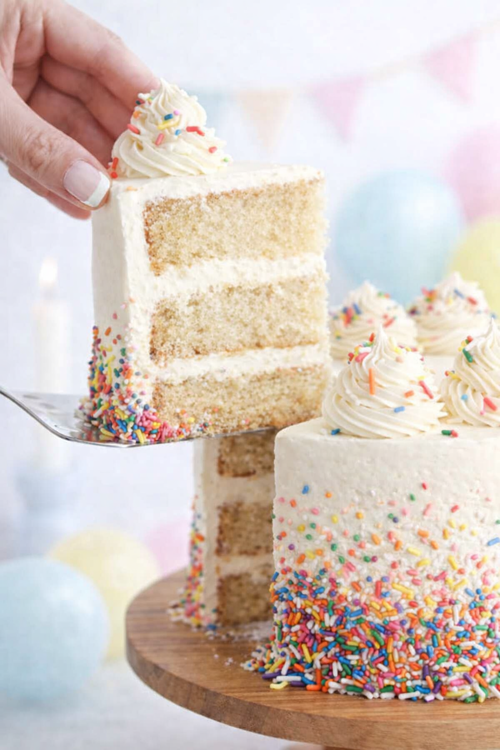Gâteau vanille à trois étages recouvert d’un dégradé de sprinkles colorés, avec une part soulevée sur une pelle à gâteau.