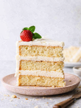 Tranche de layer cake crème vanille sur une assiette en grès rose pâle, décorée d’une fraise fraîche et d’une feuille de menthe, avec une crème légèrement imparfaite et quelques miettes visibles sur la table.