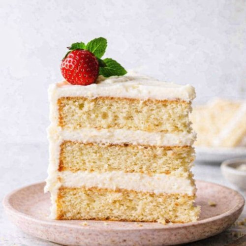 Tranche de layer cake crème vanille sur une assiette en grès rose pâle, décorée d’une fraise fraîche et d’une feuille de menthe, avec une crème légèrement imparfaite et quelques miettes visibles sur la table.
