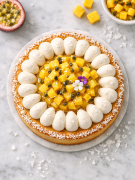 Tarte mangue, coco et passion sur assiette gris anthracite, décorée de dômes de crème coco et de dés de mangue fraîche, sur table en bois clair.